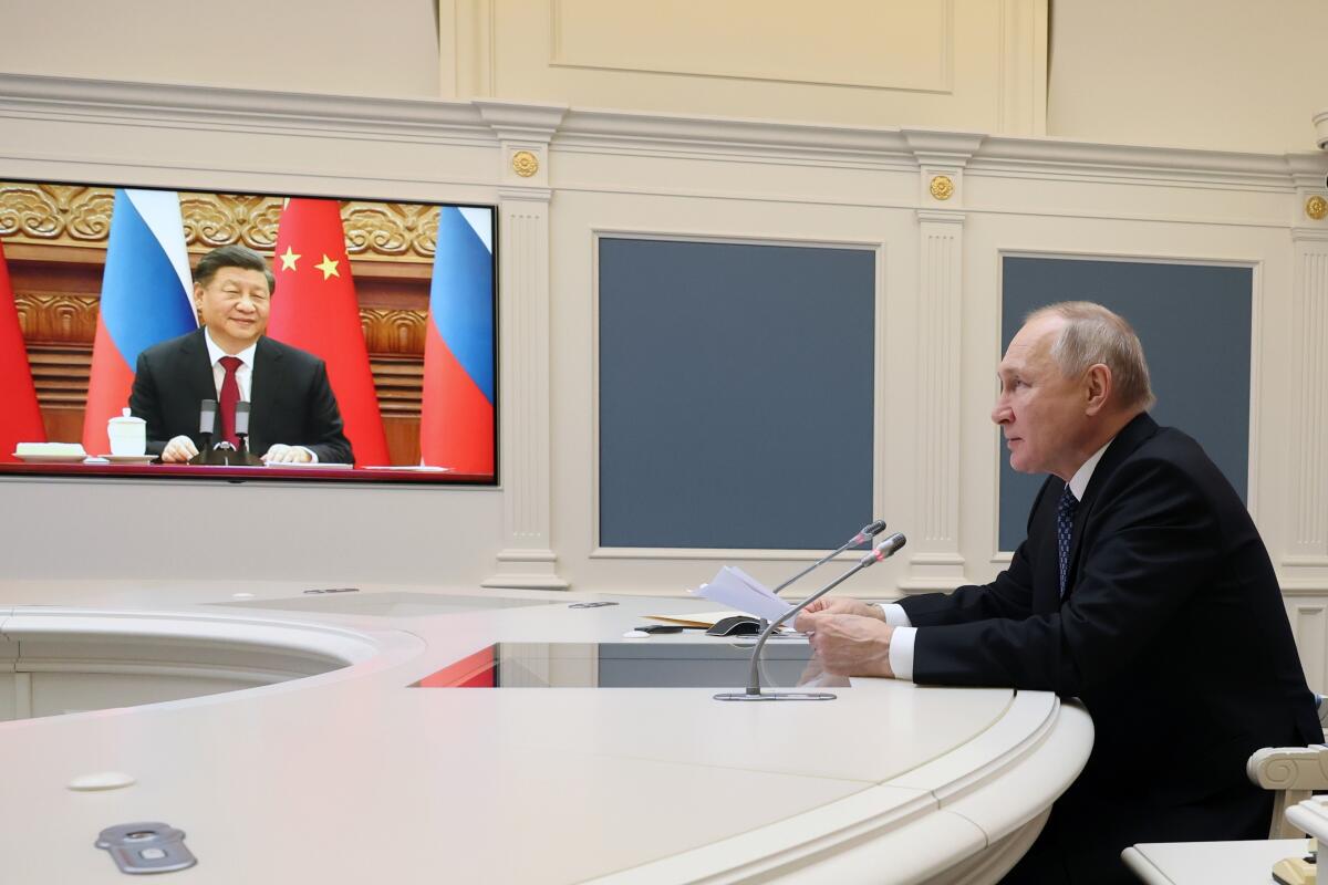 A man sits alone at a conference table overlooked by a video screen with the image of another man sitting in front of flags