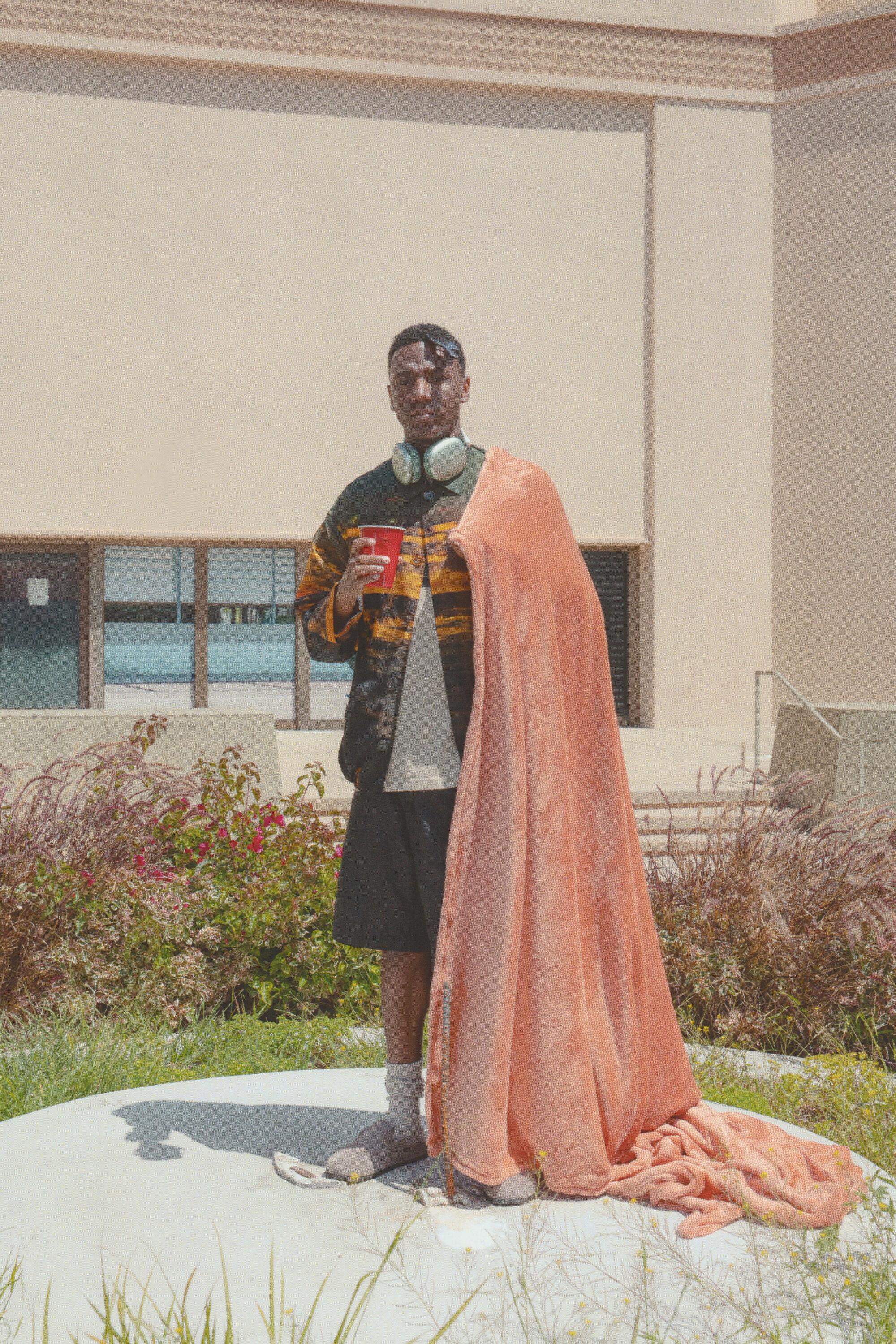 A man with headphones around his neck and a red solo cup in his right hand and a fleece blanket draped over his shoulder