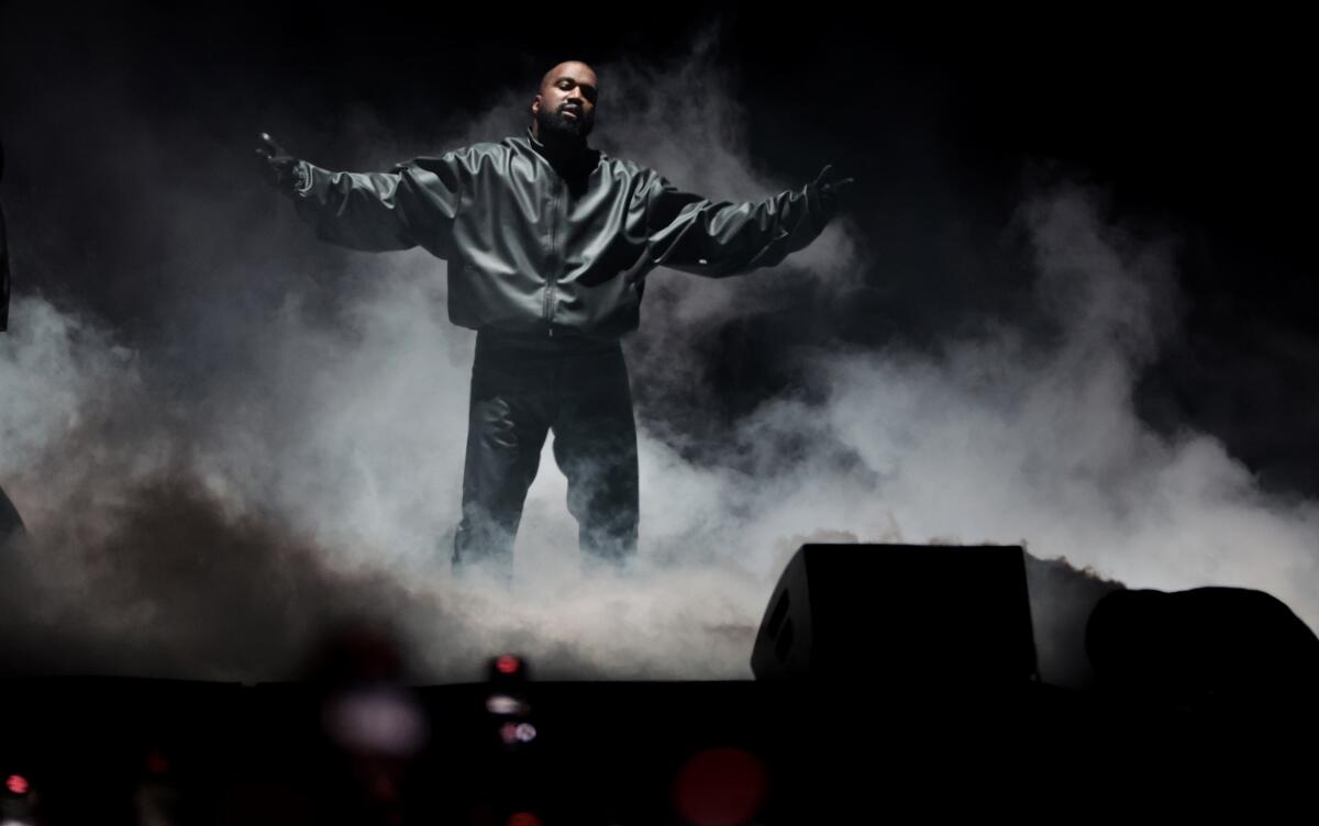 Kanye West in a black jacket, black pants standing on a foggy stage