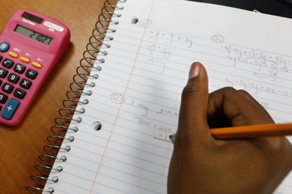 A student's hand writes math work in a notebook next to a calculator