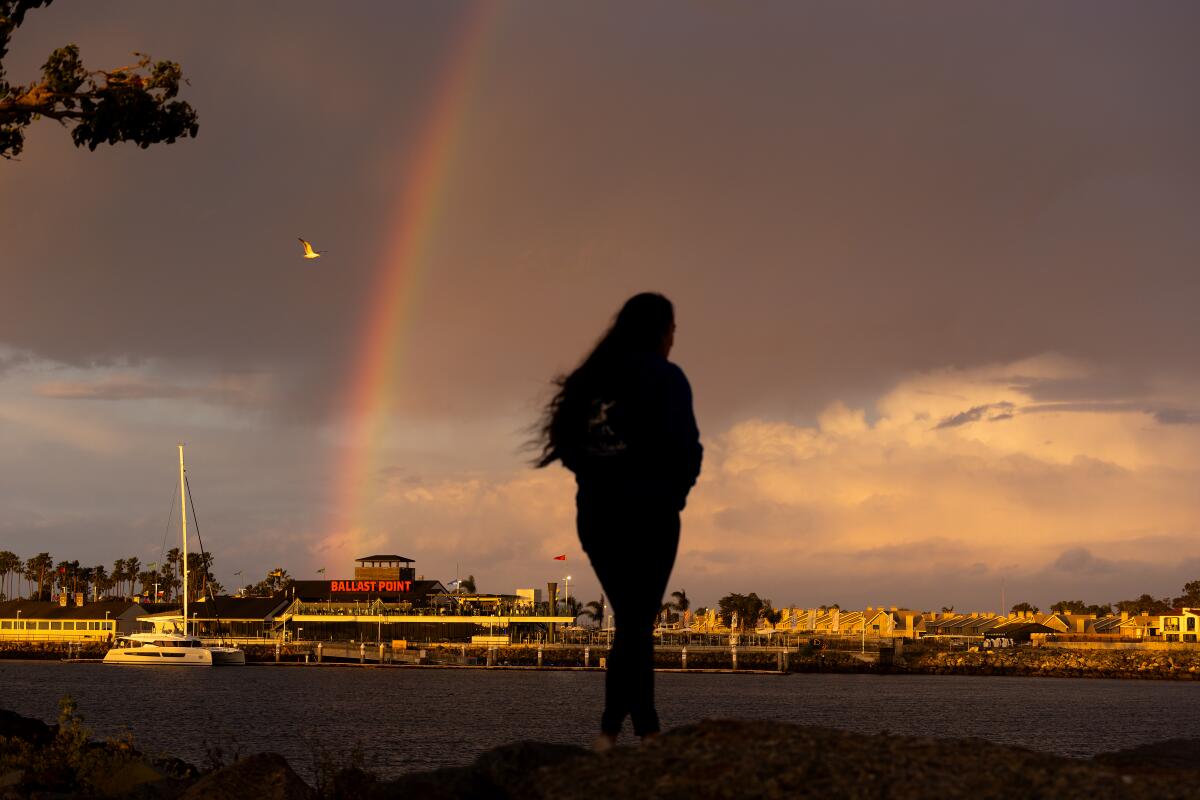 A rainbow appears over Ballast Point Brewing Long Beach amid light rain as a person in silhousette walks in the foreground