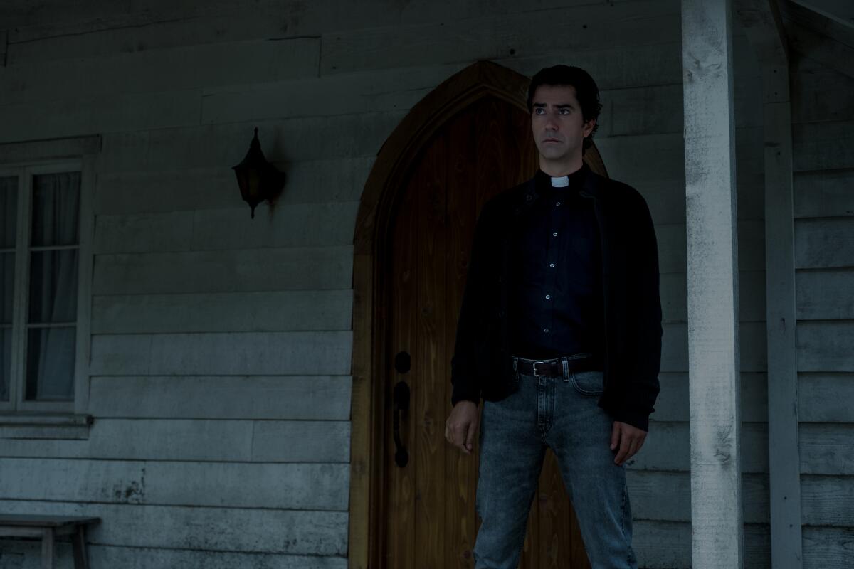 Hamish Linklater as Father Paul stands on a porch in priest's collar and jeans.