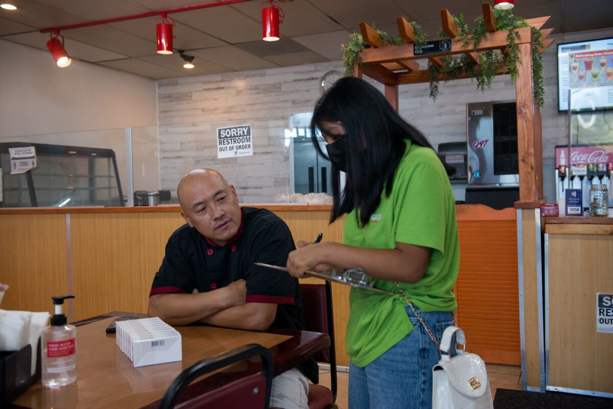 A teen with a clipboard speaks to a restaurant worker sitting at a table