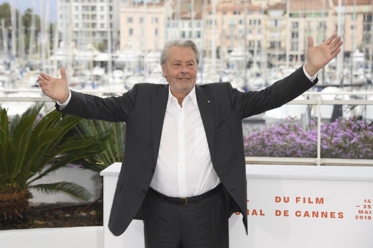 Alain Delon in a dark suit and white shirt posing in front of sailboats and buildings with his arms open wide