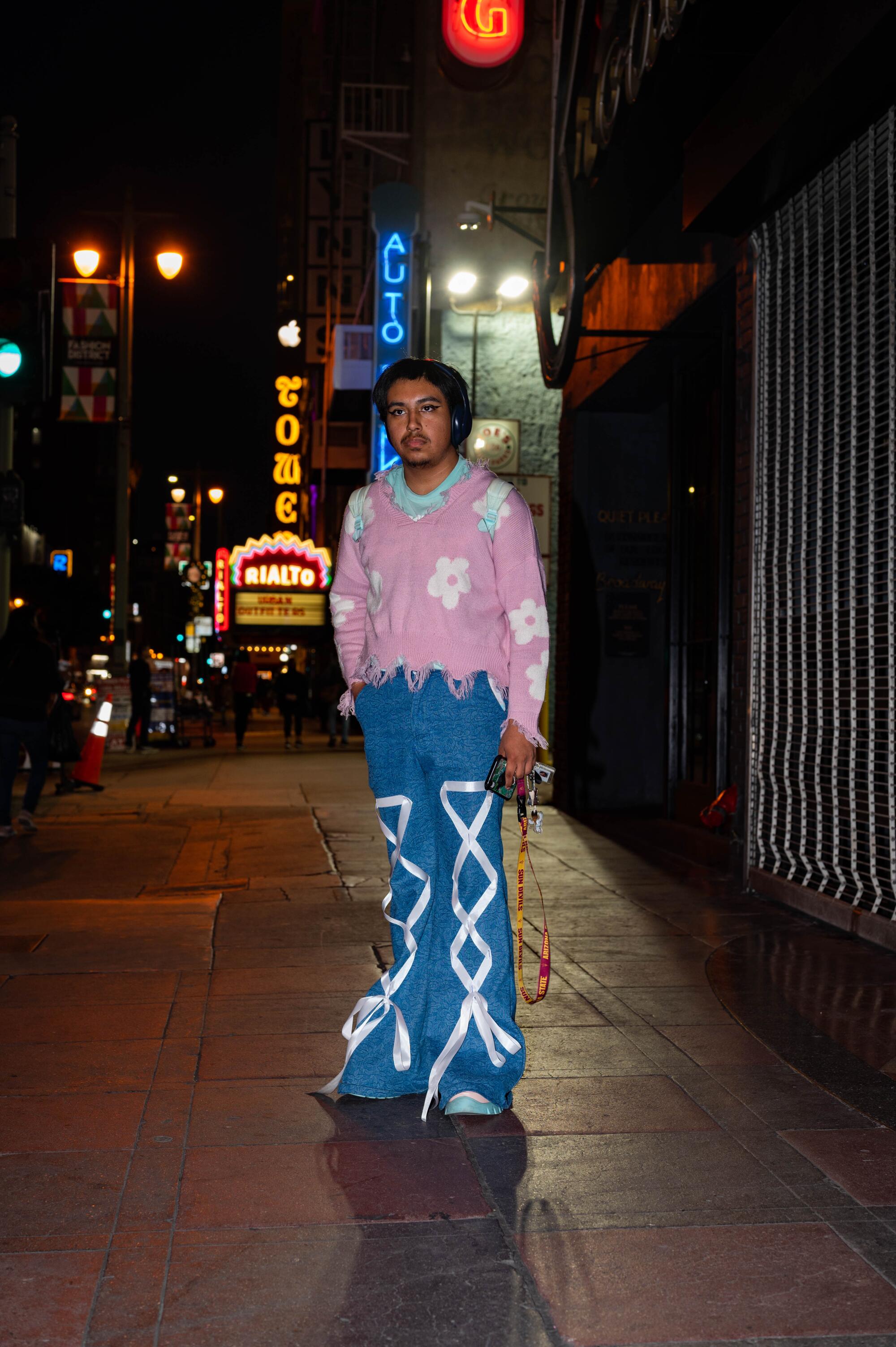 A person in blue pants and a pink blouse is standing on the sidewalk, with neon signs behind them.