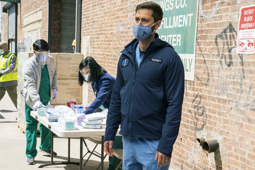 A man in scrubs and a face mask stands on the street near a table where workers sort medical equipment.