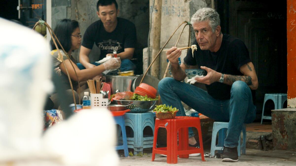 Anthony Bourdain eats with chopsticks sitting on a low stool.