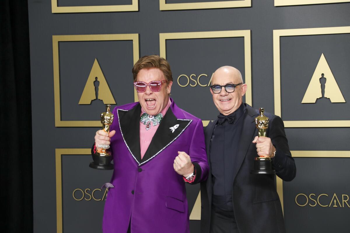 Elton John in a purple tuxedo and Bernie Taupin hold Oscars