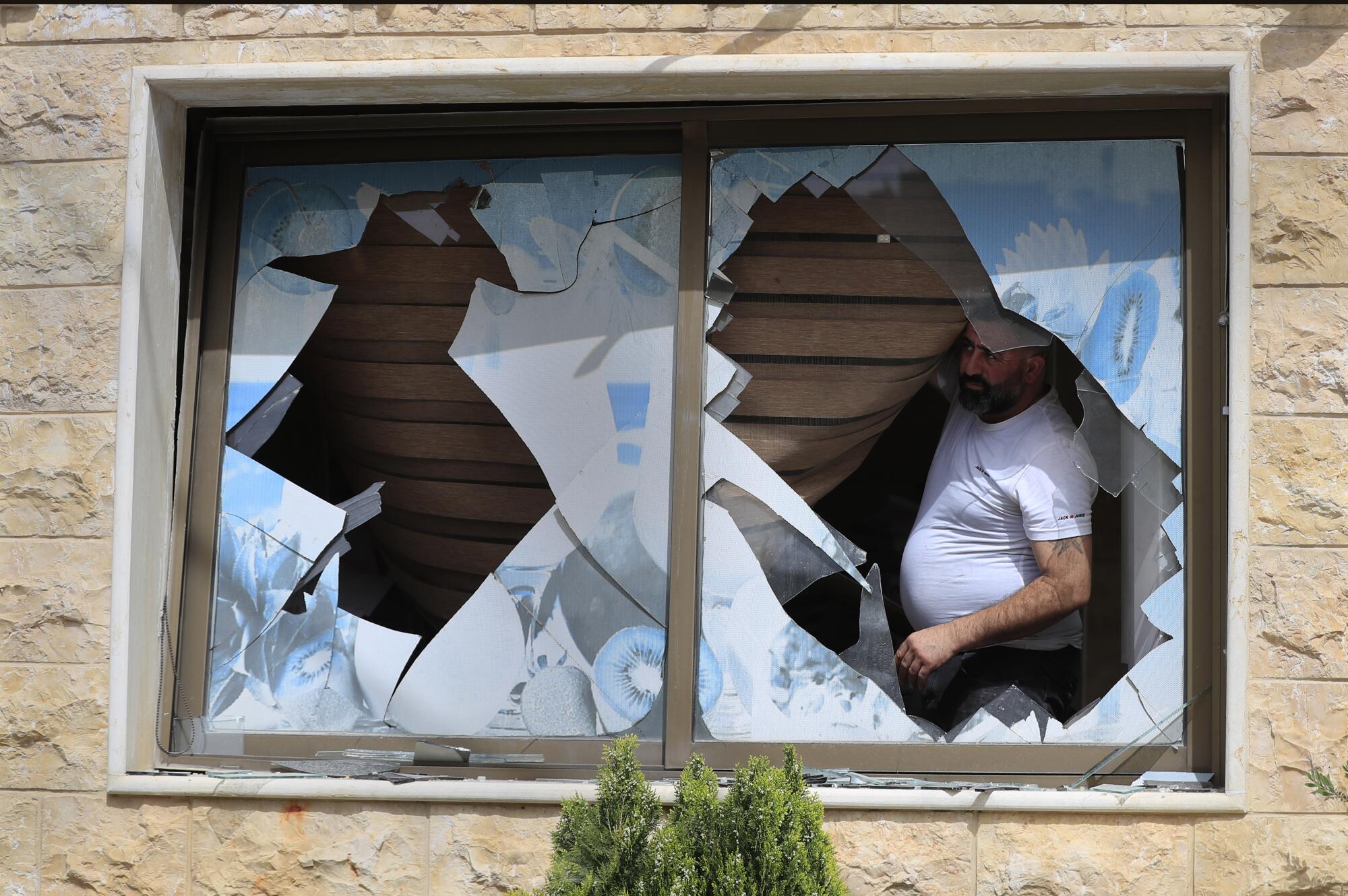 A man in a white T-shirt looks through the jagged opening of his broken window. 