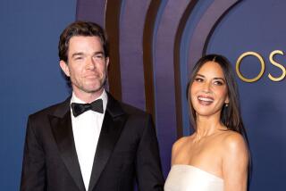 John Mulaney in a tuxedo and Olivia Munn in a strapless white gown smiling as they arrive together at the Oscars