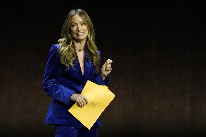A woman in a blue dress and holding a large envelope speaks to an audience.