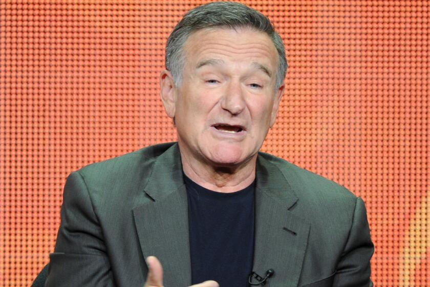 Robin Williams opens his mouth and gestures with his hands while sitting in front of an orange background