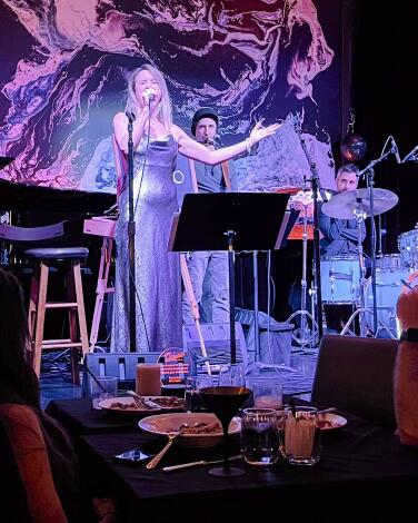A woman sings onstage in a club, with diners at a table next to the stage