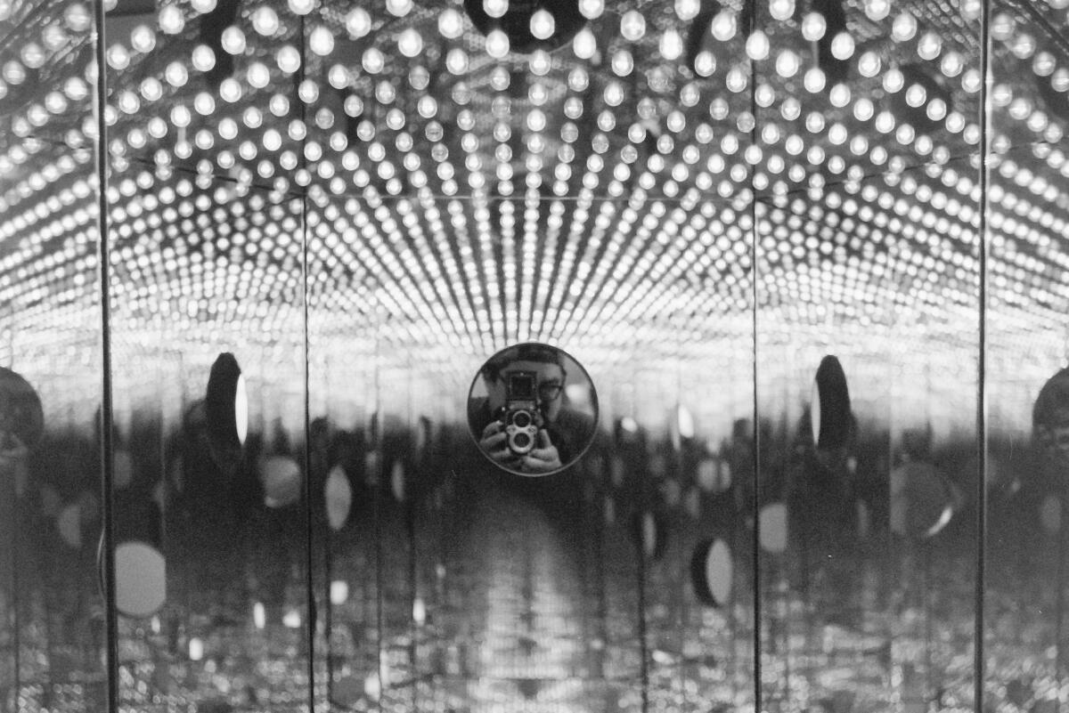 A man with glasses, looking through a camera lens, is at the center of a mirrored surface reflecting hundreds of lights. 