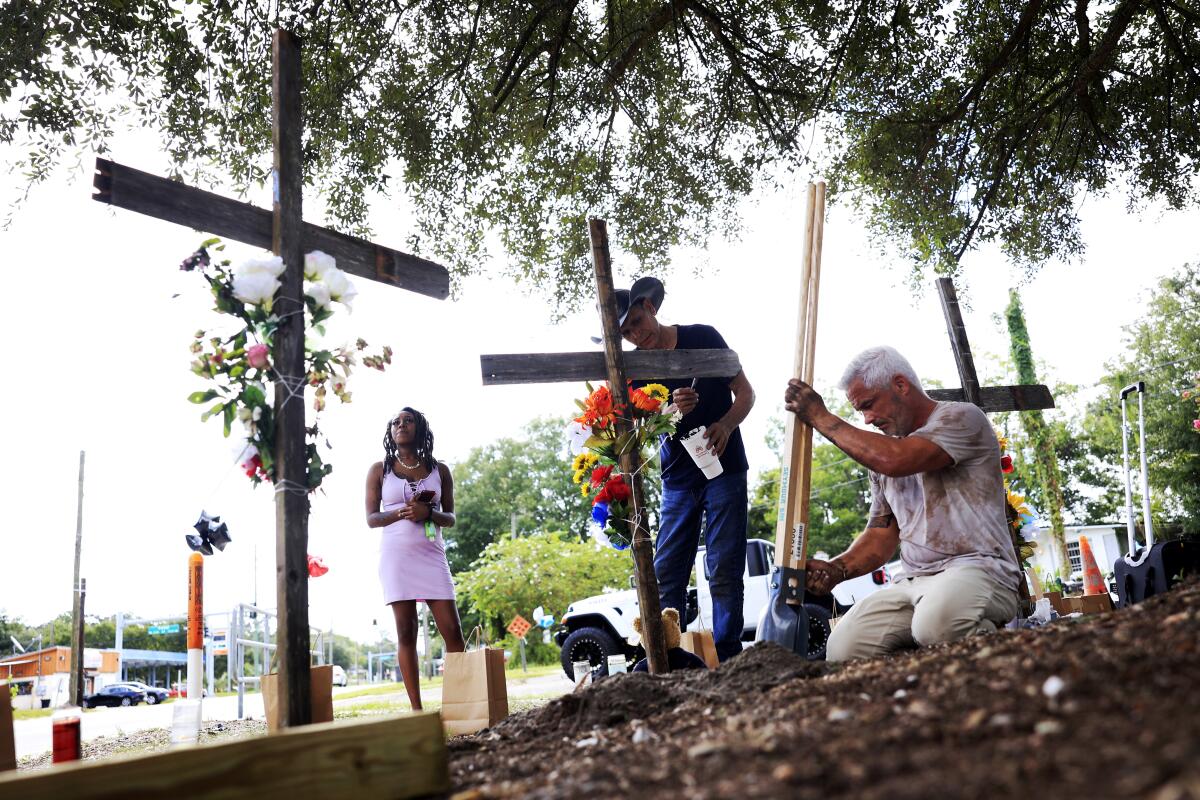 People look at crosses adorned with flowers in the shade of trees 