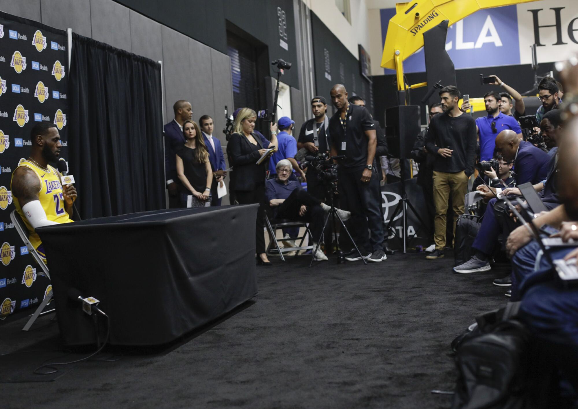 LeBron James, in Lakers uniform, sits behind a table holding a microphone. Media members are packed nearby. 