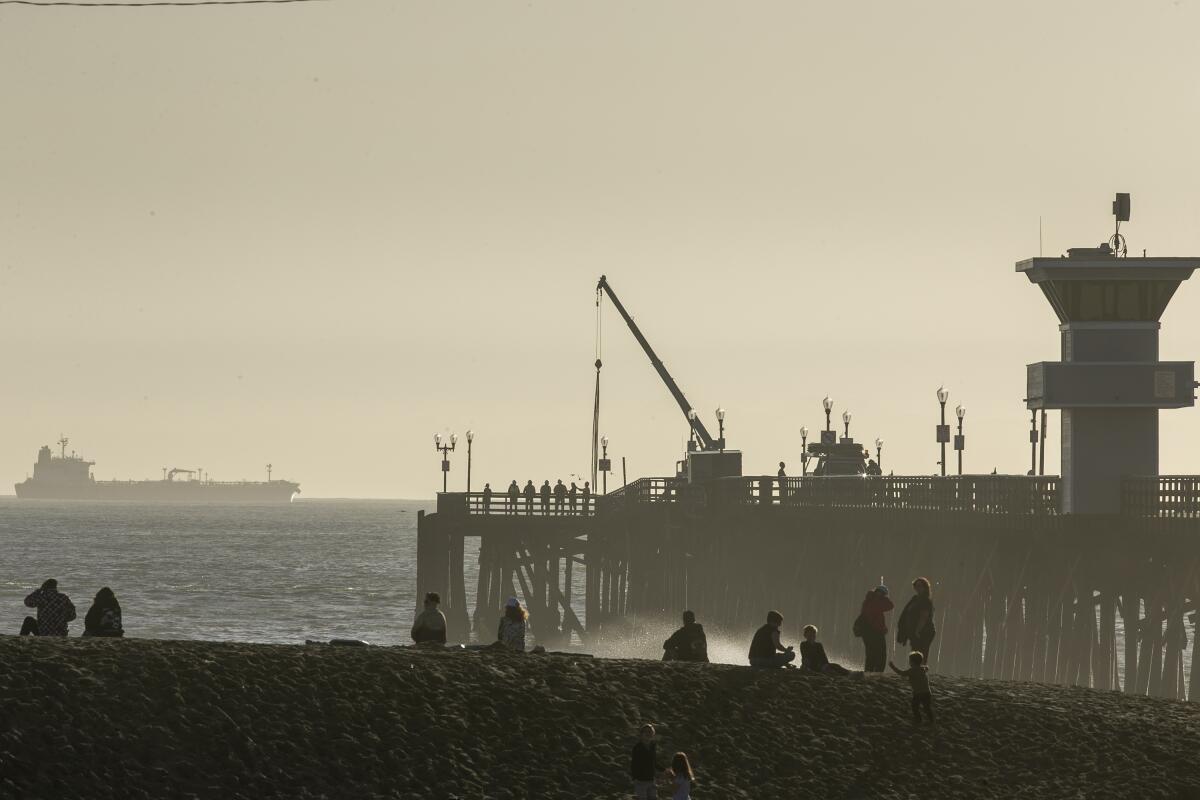 Crews Friday work on the Seal Beach Pier, damaged by winds and surf Thursday night, while beachgoers sit on a sand berm.
