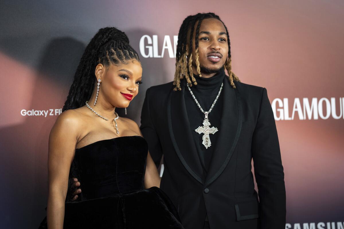 Halle Bailey and DDG, both in black formalwear, pose with arms around each other's waists in front of a backdrop