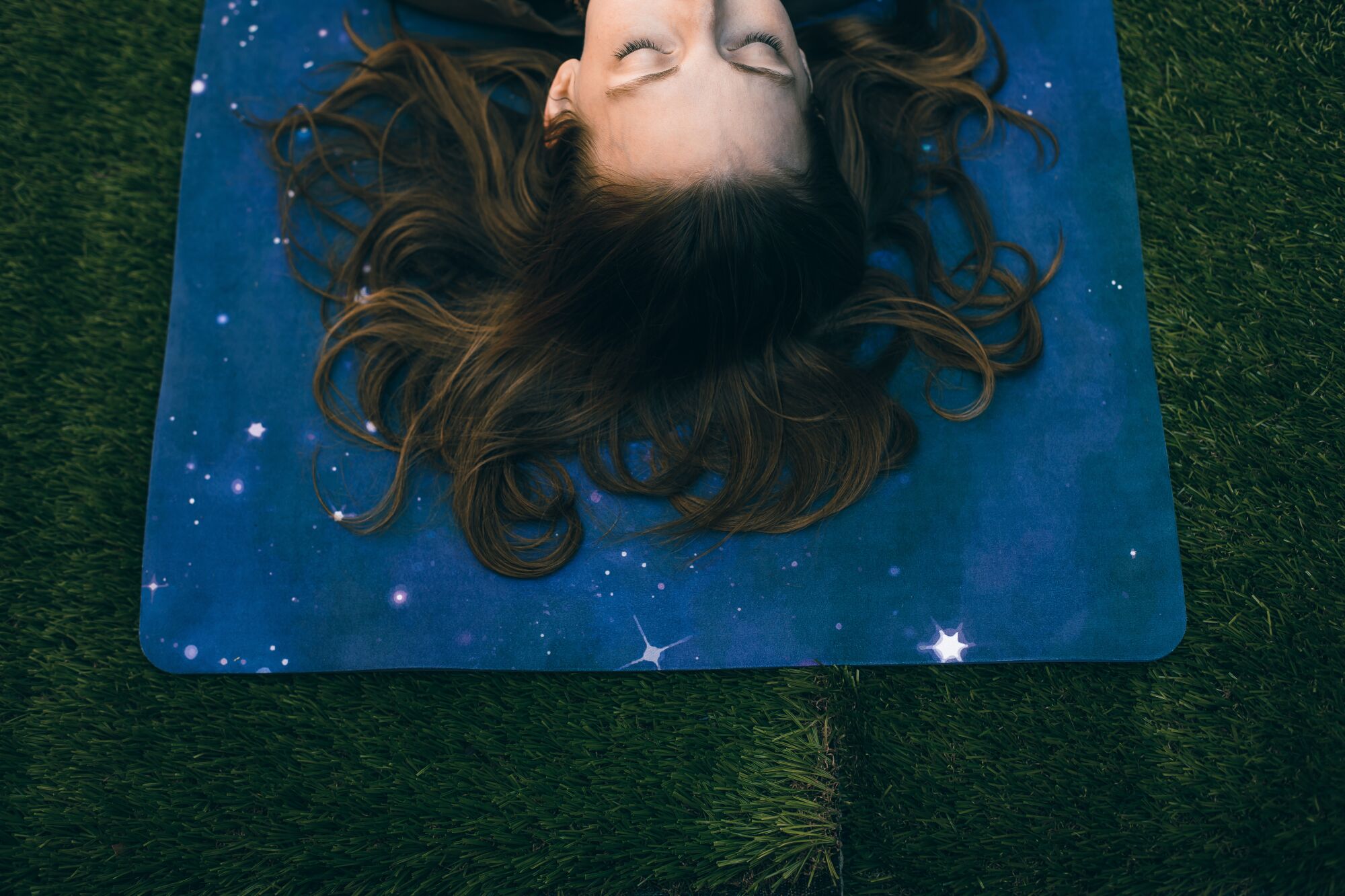 A view from above of a person lying on a galaxy-themed yoga mat on the grass, only their hair and top of their face visible