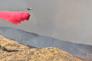 A pink cloud is seen trailing behind an aircraft flying over smoky hills.