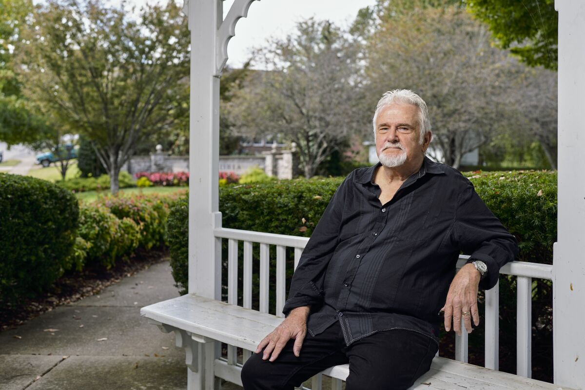A man with white hair and a goatee sits on a bench outdoors with a garden behind him.