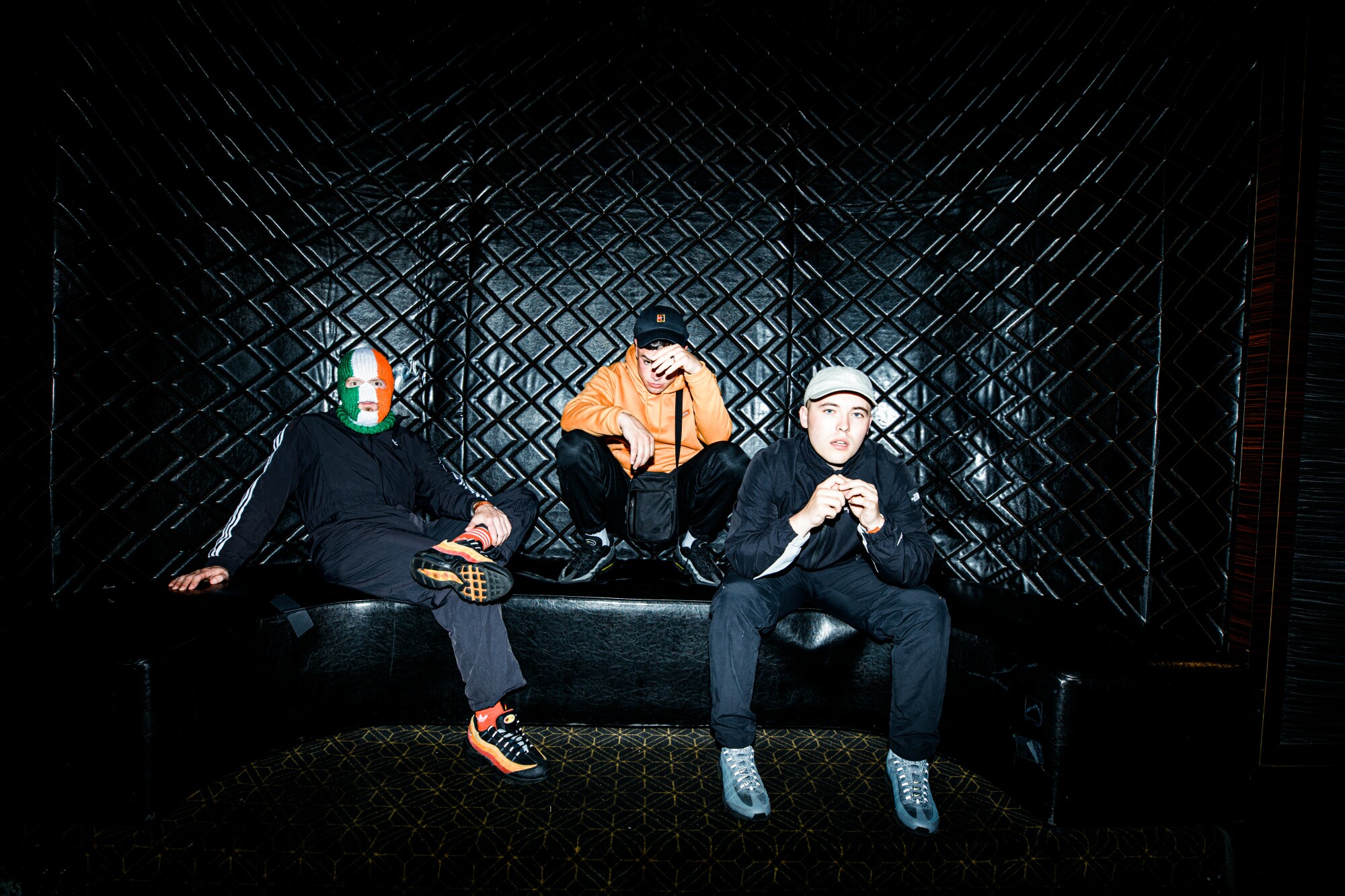 Three men sit in front of a black wall with a diamond pattern.