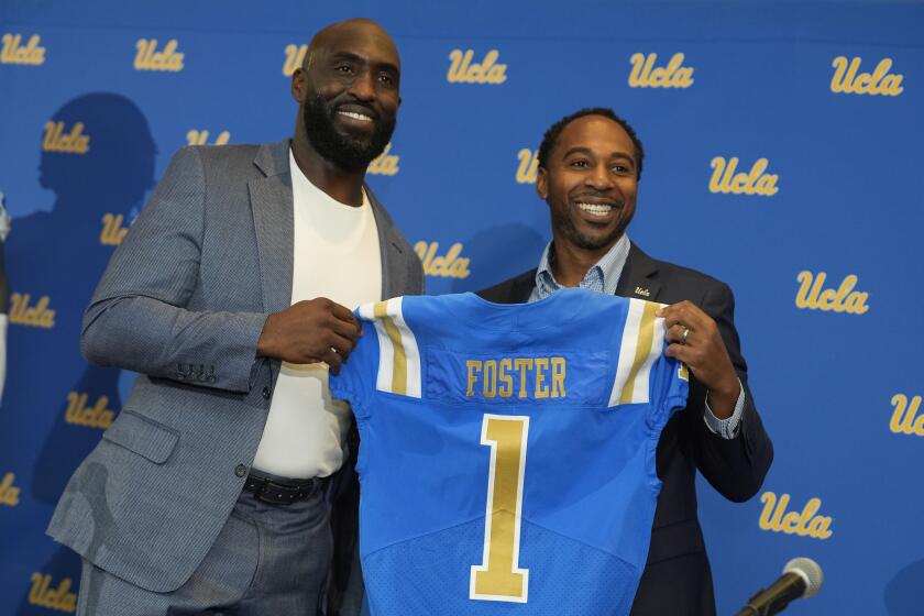 New UCLA head football coach DeShaun Foster holds up a jersey and poses alongside athletic director Martin Jarmond