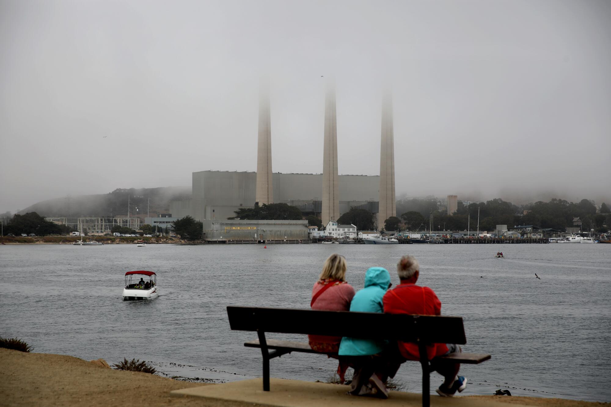 Three people sit on a bench overlooking the sea with a power plant and three smokestacks in the background