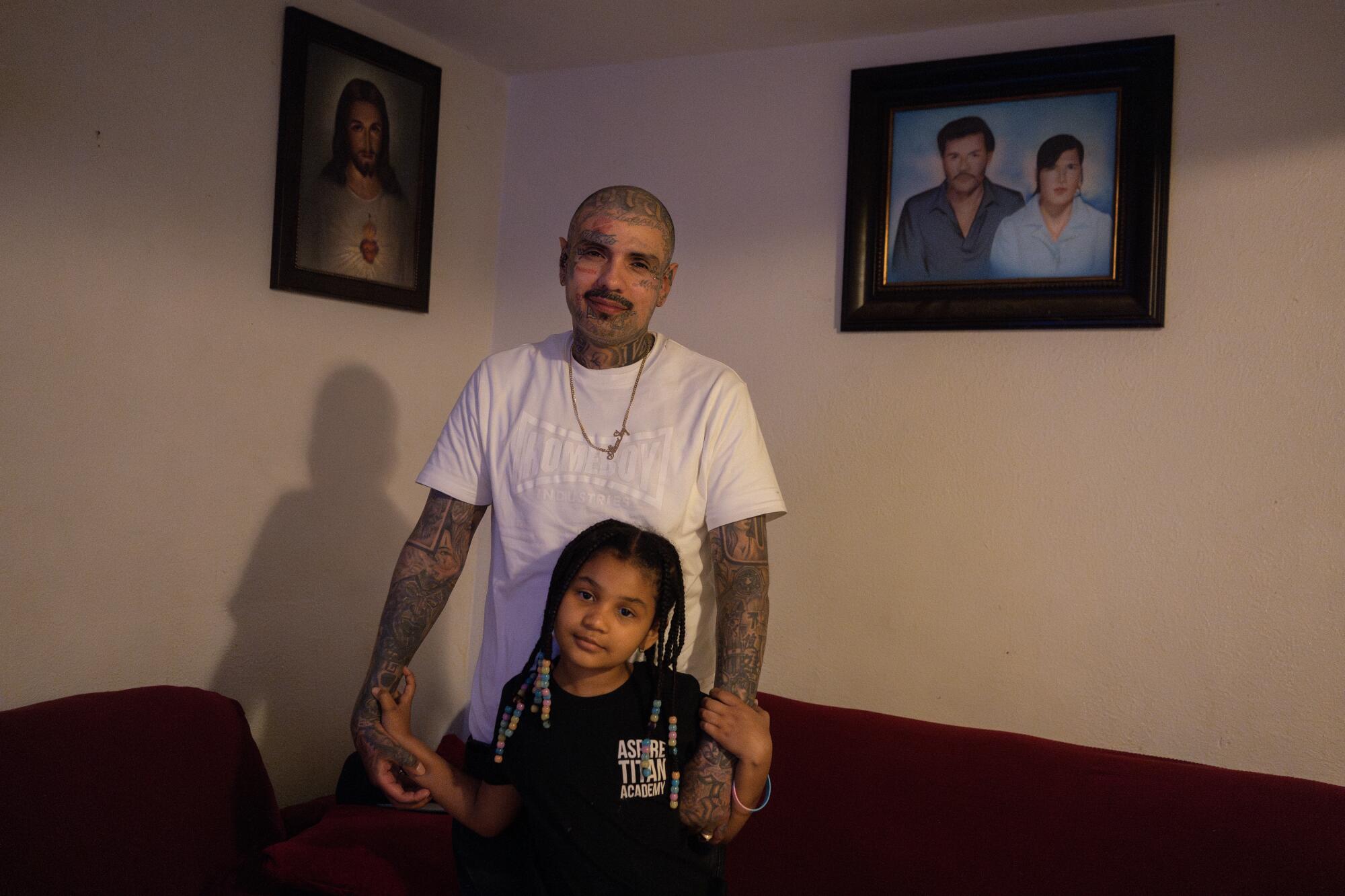 A man in a white T-shirt and covered by tattoos and a girl with braided hair hold hands and face the camera.