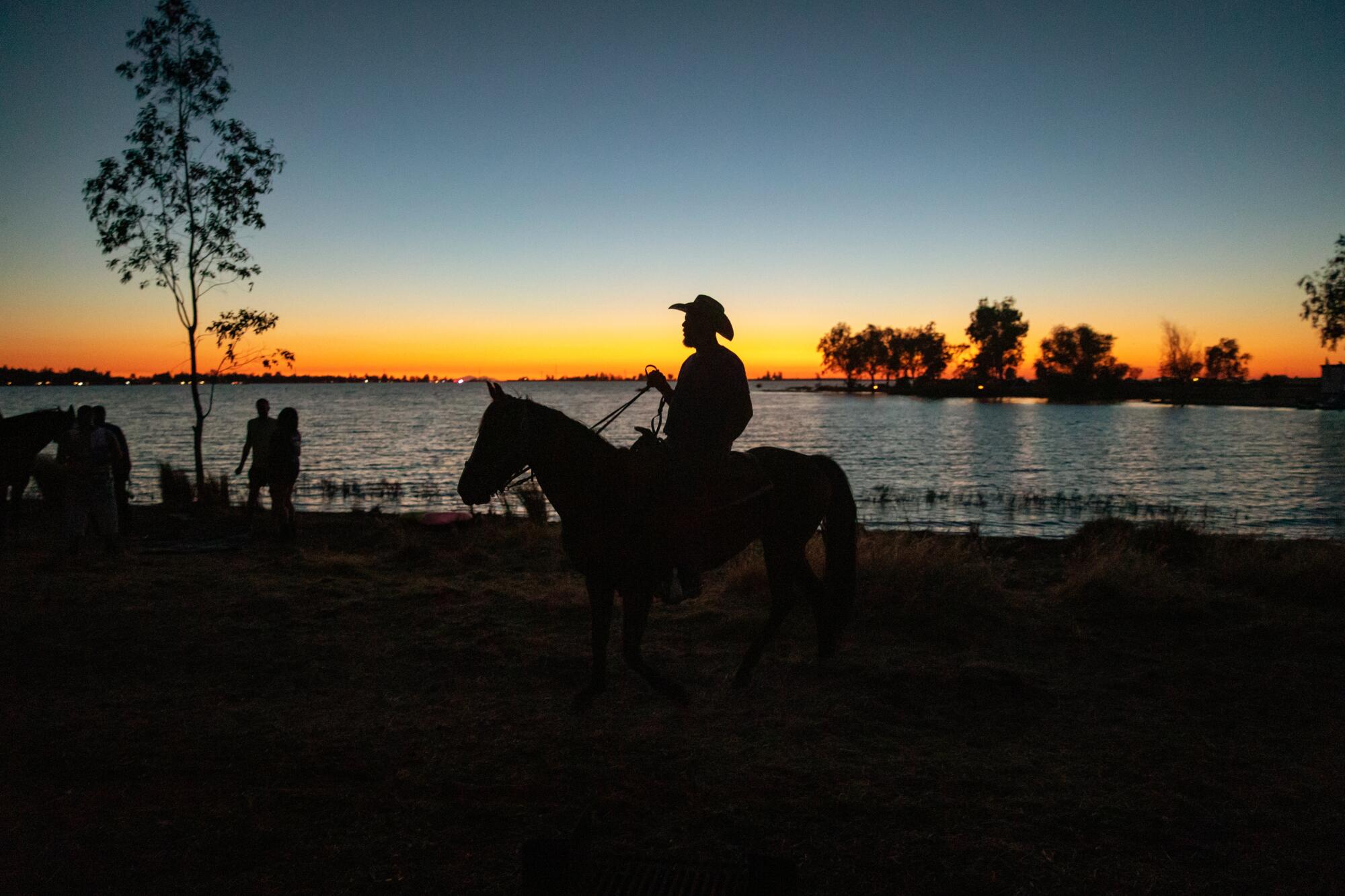 The silhouette of a cowboy on a horse against a body of water at sunset.