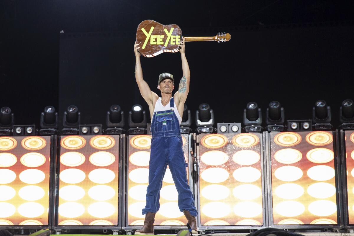 Granger Smith, in overalls, holds a guitar over his head.