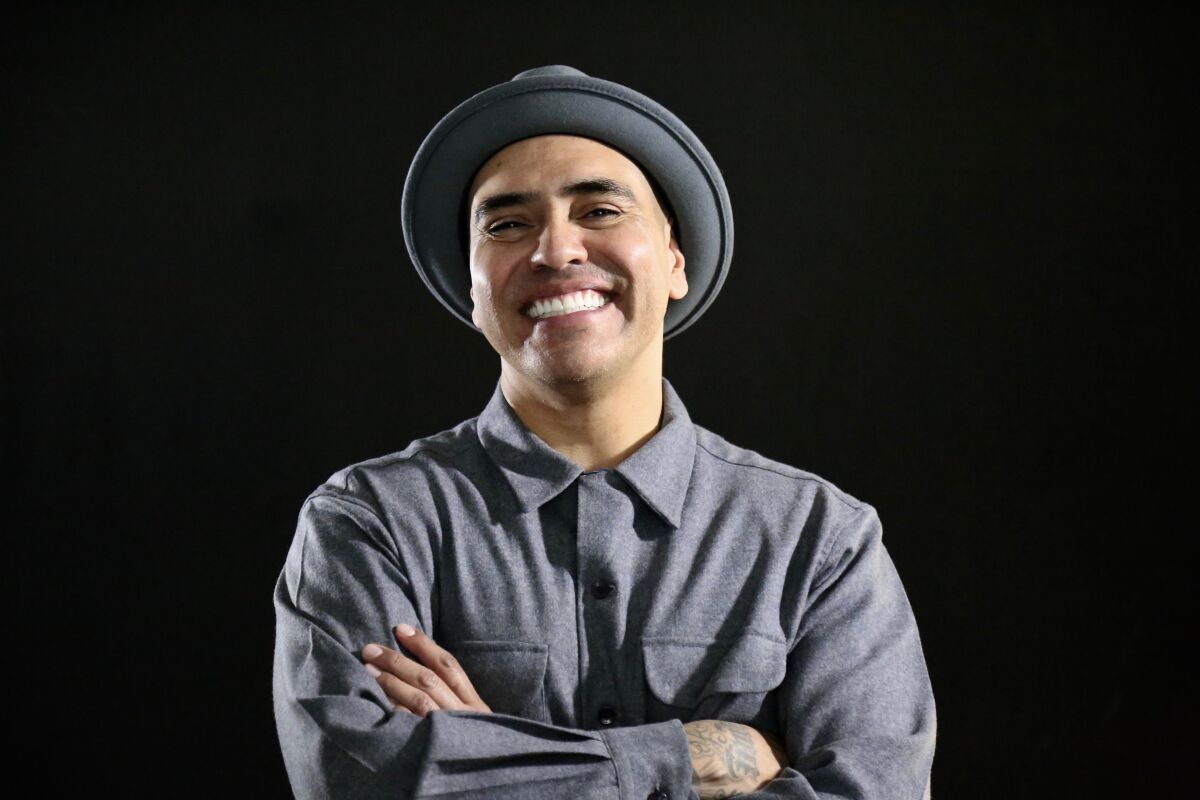 A smiling man wearing a gray hat and gray long-sleeved shirt stands with arms crossed.