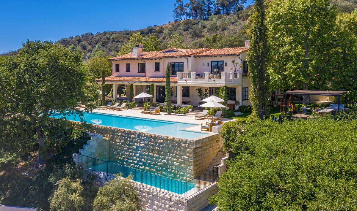 Swimming pools on two levels sit behind a two-story mansion on a tree-covered hillside.