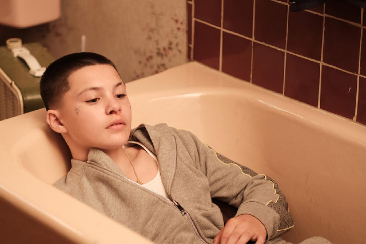 A young boy with a face tattoo lays in wait in an empty bathtub