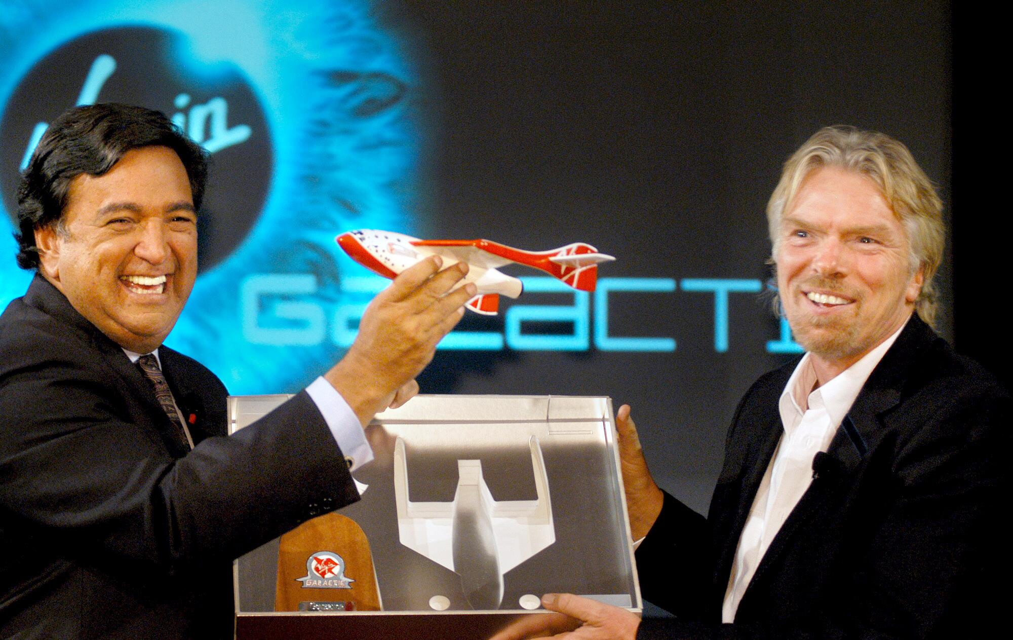 A smiling man with dark hair, left, holds a model of a spaceship next to a bearded man, also smiling, holding a display 