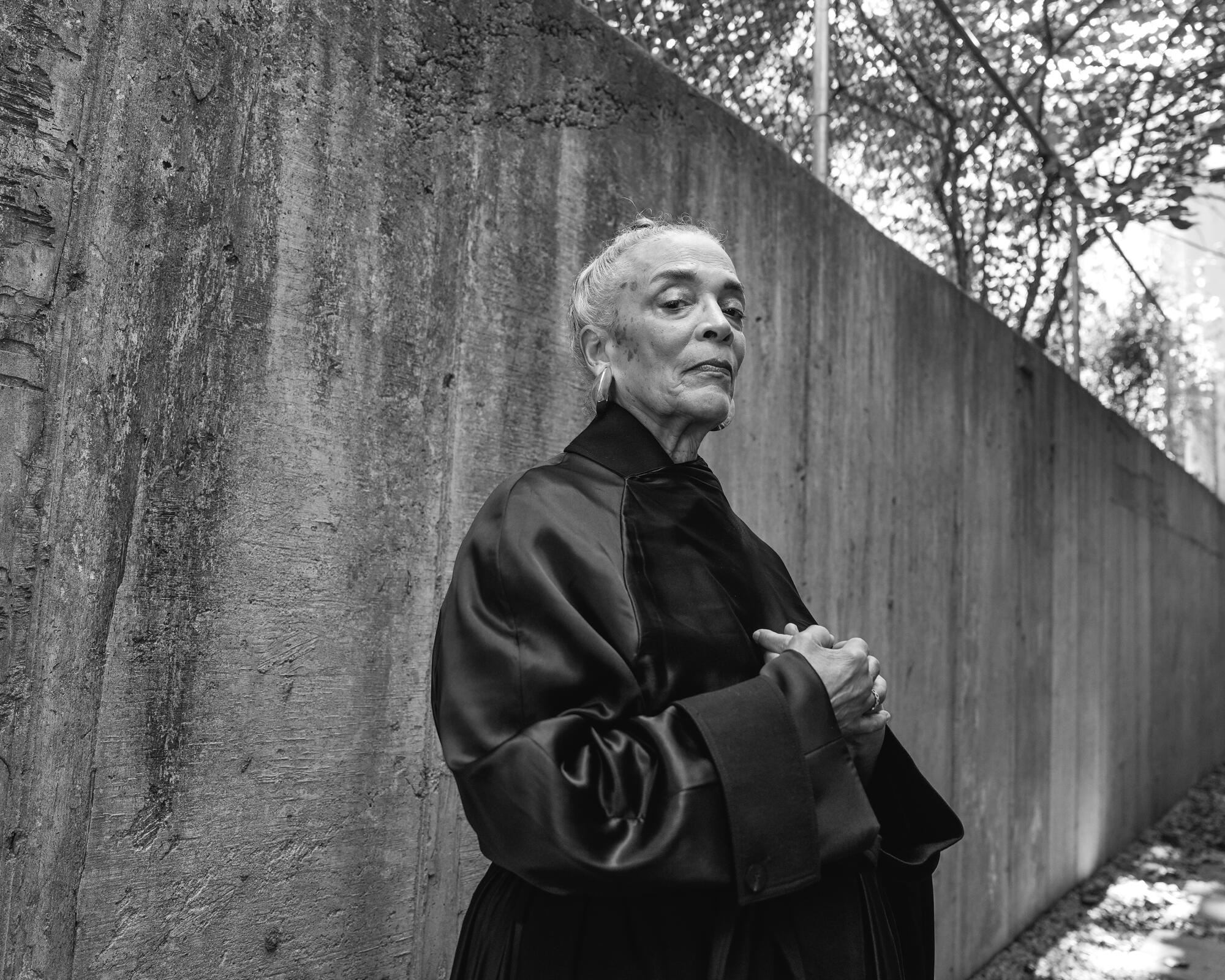 A black and white photo of a woman wearing a black robe beside an outdoor concrete wall.
