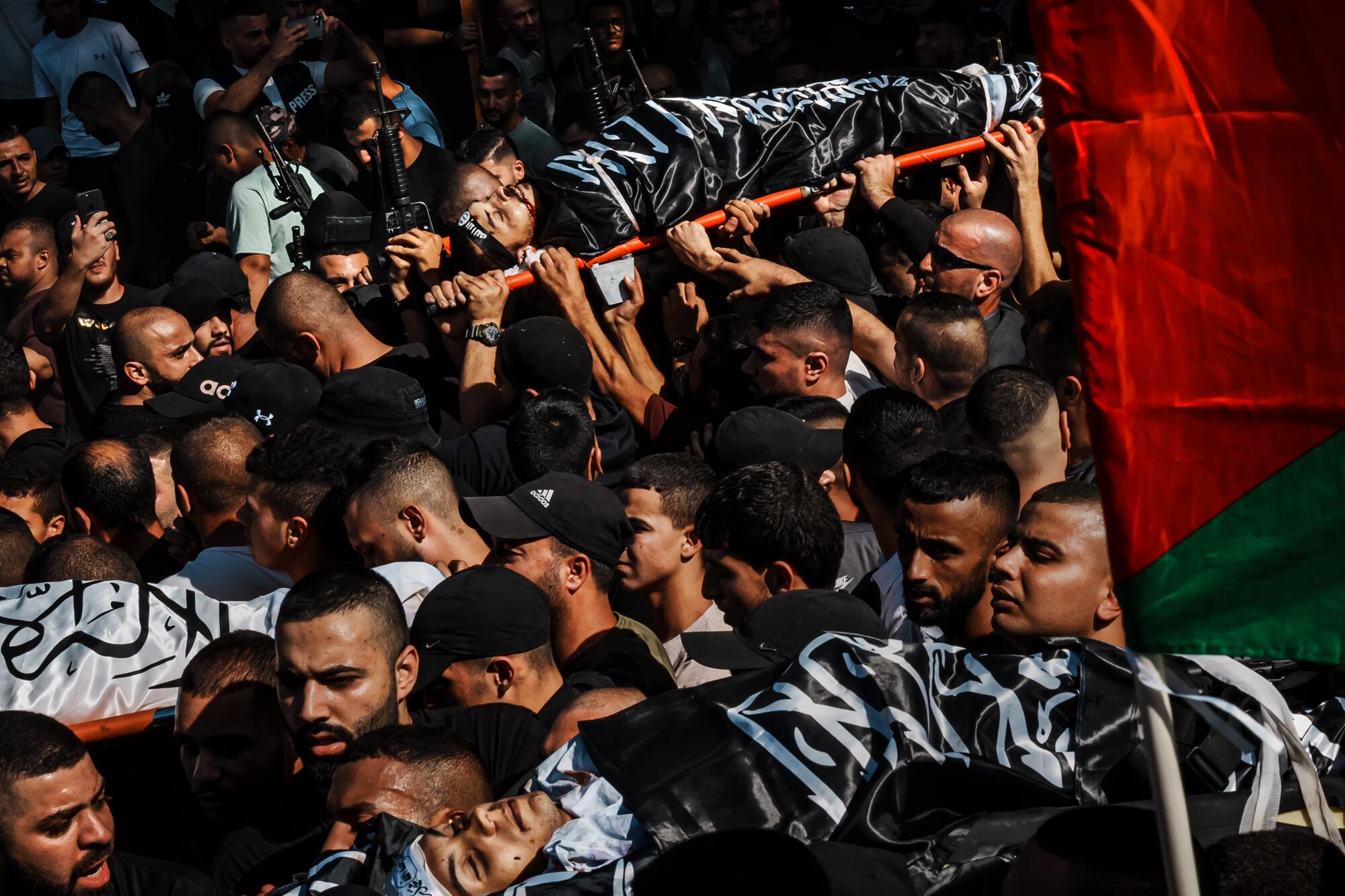 People in a crowd hold up a black-covered body on a stretcher near red-and-green flag 