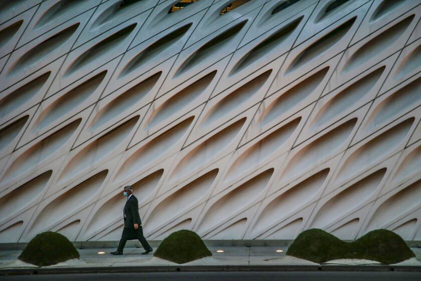 A man in a suit and a face mask walks down on a sidewalk in front of the Broad Museum