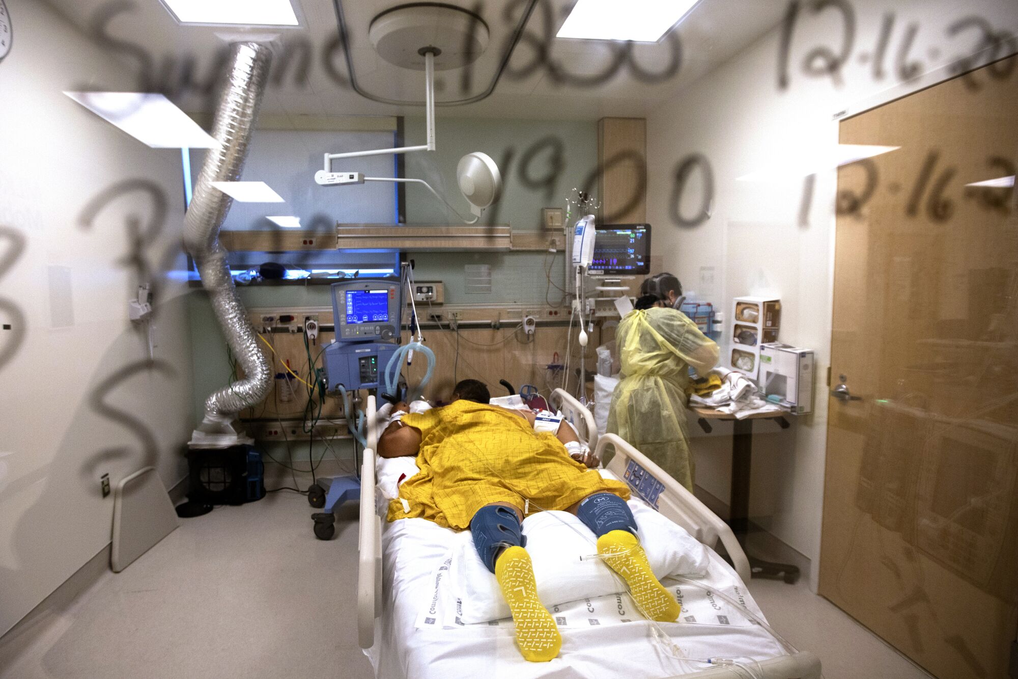 A patient lies face down on a hospital bed. A nurse in protective gear works nearby.