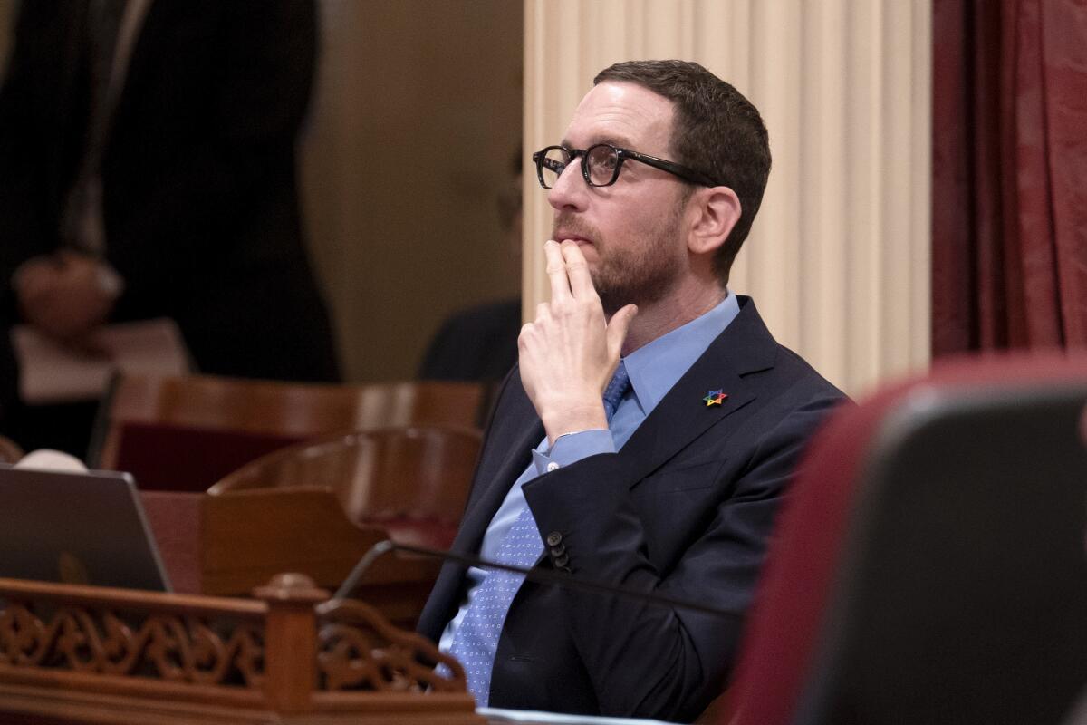 A man seated in a legislative chamber holding his hand at his face 