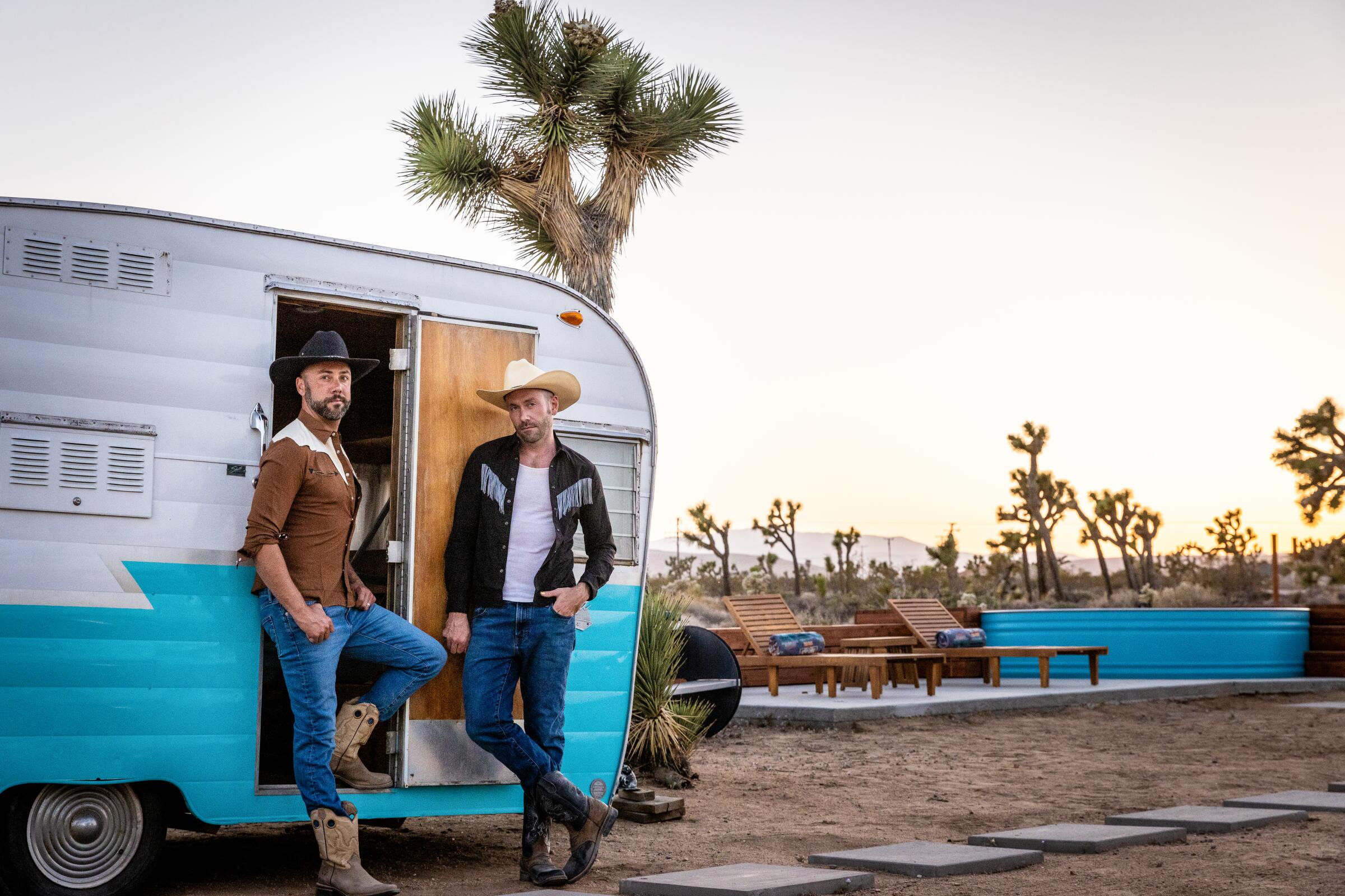 Two men in western garb and cowboy hats stand in front of a camper next to a stock tank pool.