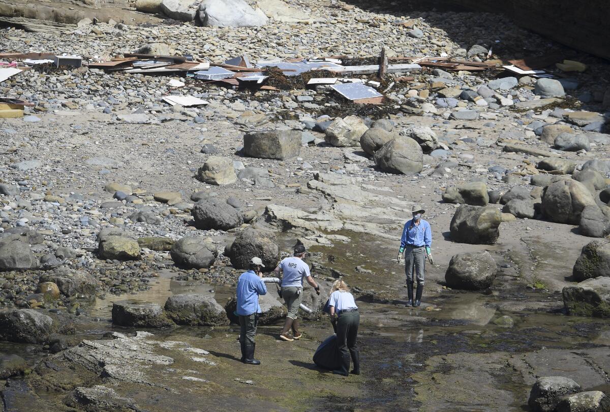 People stand on a rocky shoreline next to debris from a wrecked boat