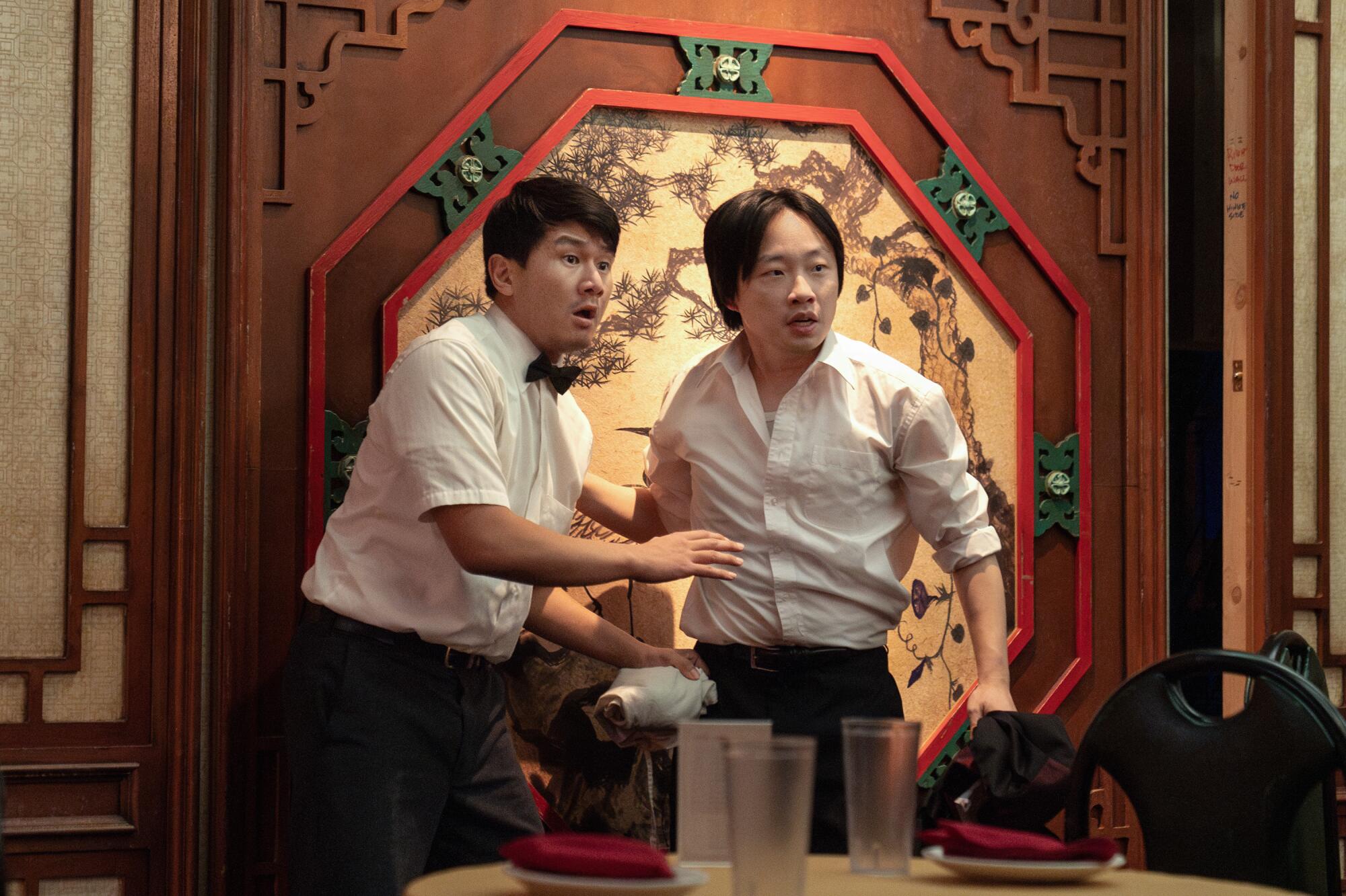 Two alarmed waiters in white shirts stand in front of a dining table.