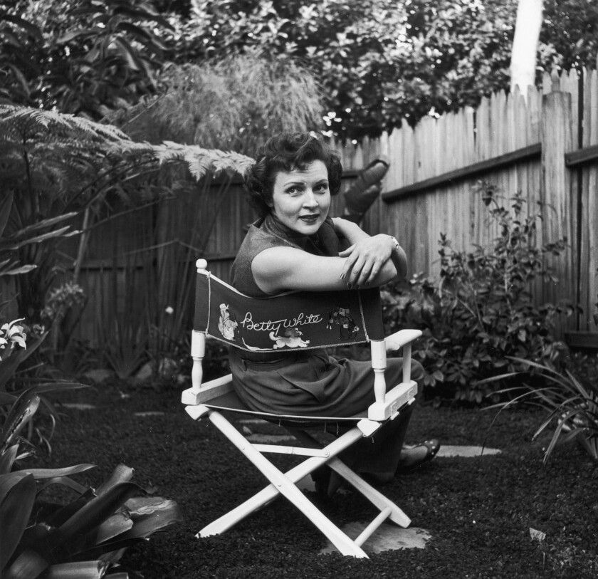 Betty White sits in a canvas chair with her name written on the back, looking over her shoulder in a backyard garden.