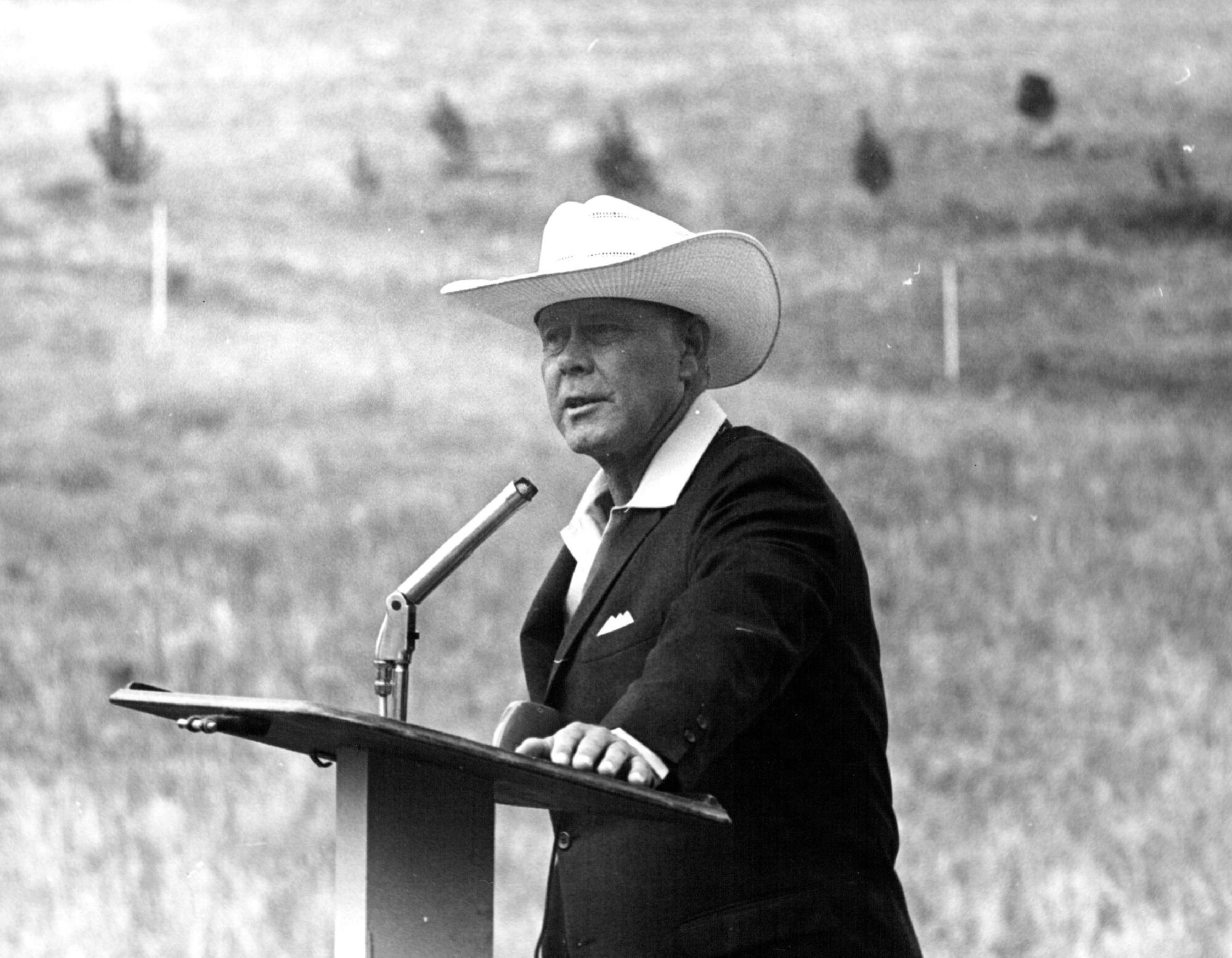 Black and white image shows man in light cowboy had and dark suit at a lectern outdoors. 