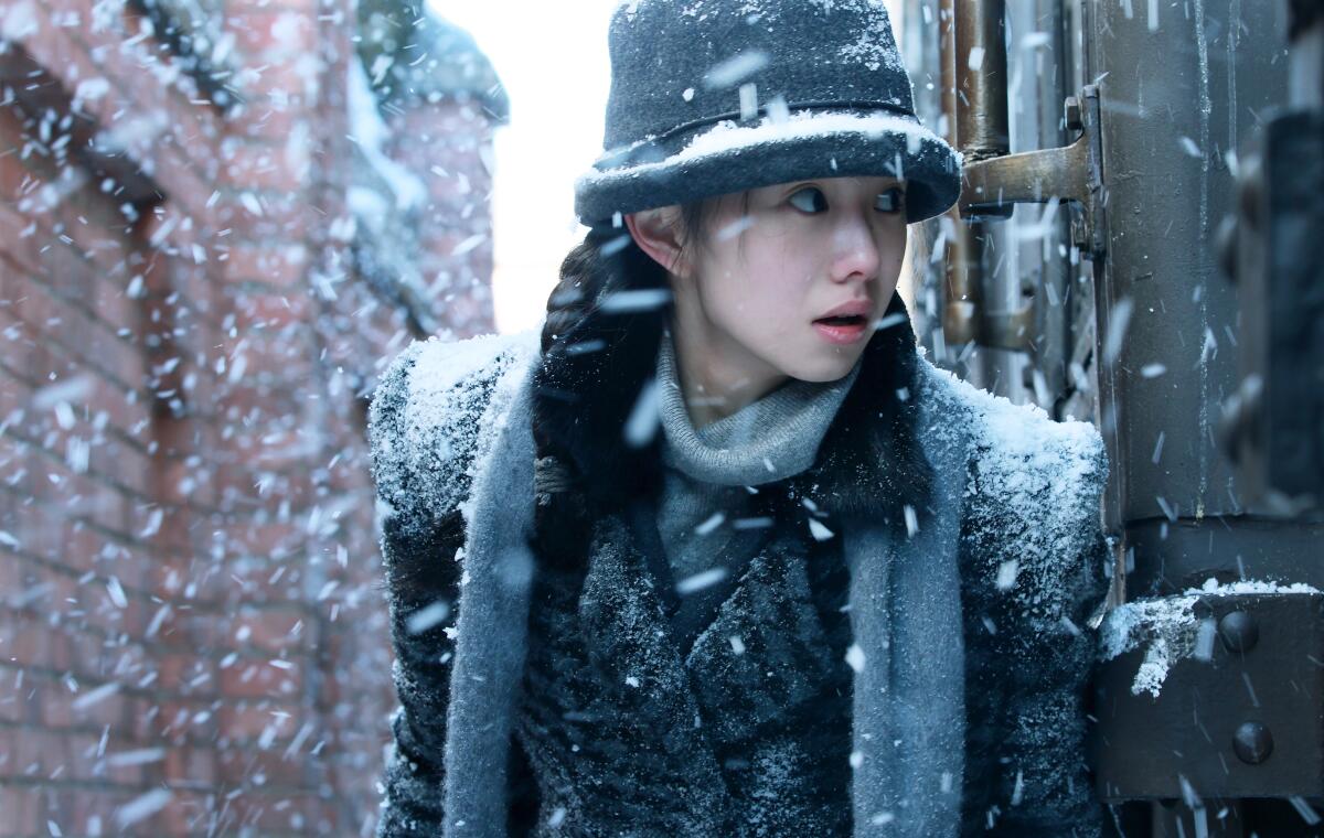 Liu Haocun, in winter coat and hat, walks amid falling snow near a brick building.