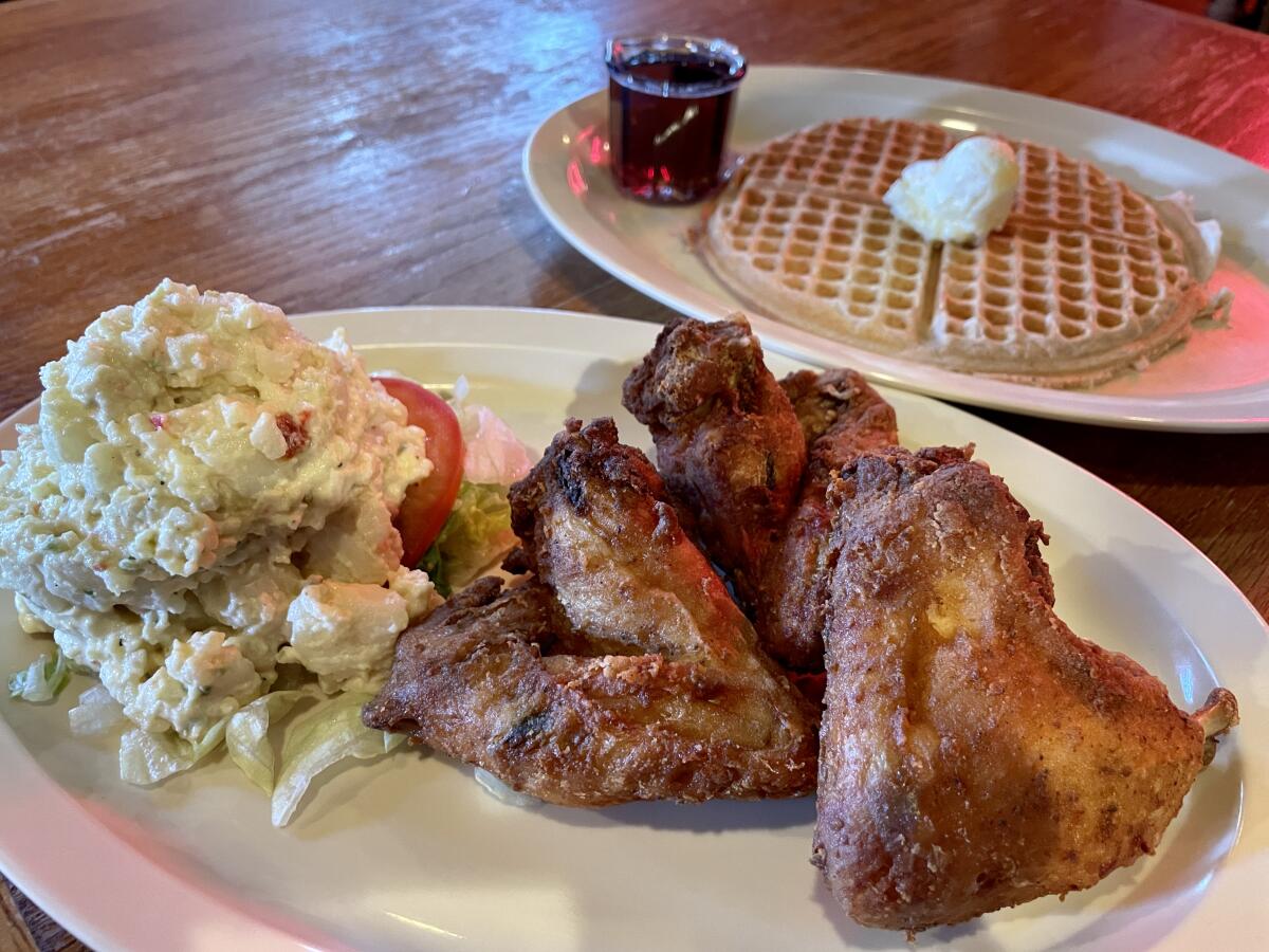 Two plates, one with chicken wings and potato salad, the other with a waffle with butter and syrup.