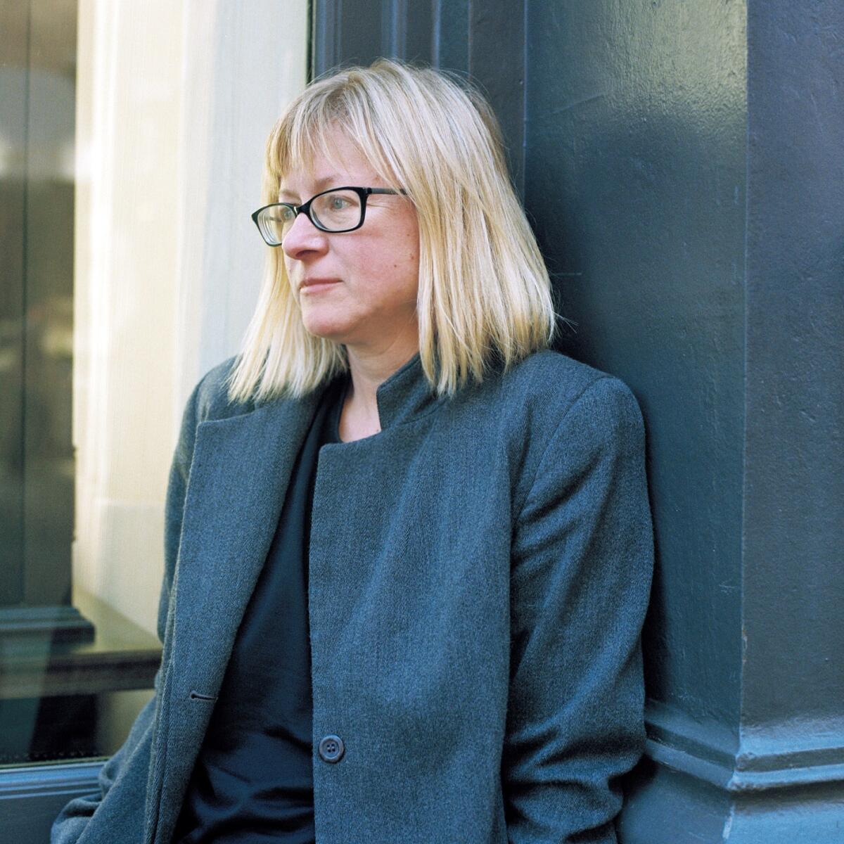 A blond woman with black glasses and a gray coat leans against a wall.