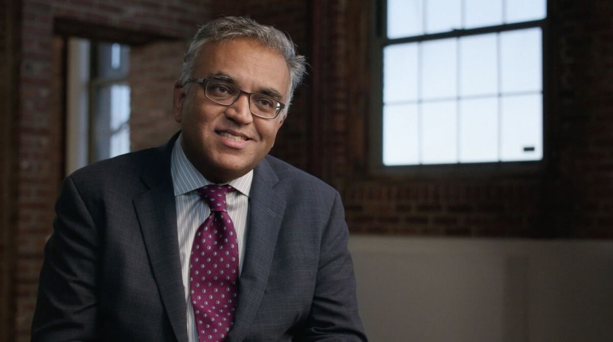 A man in a gray suit sitting in a room with brick walls