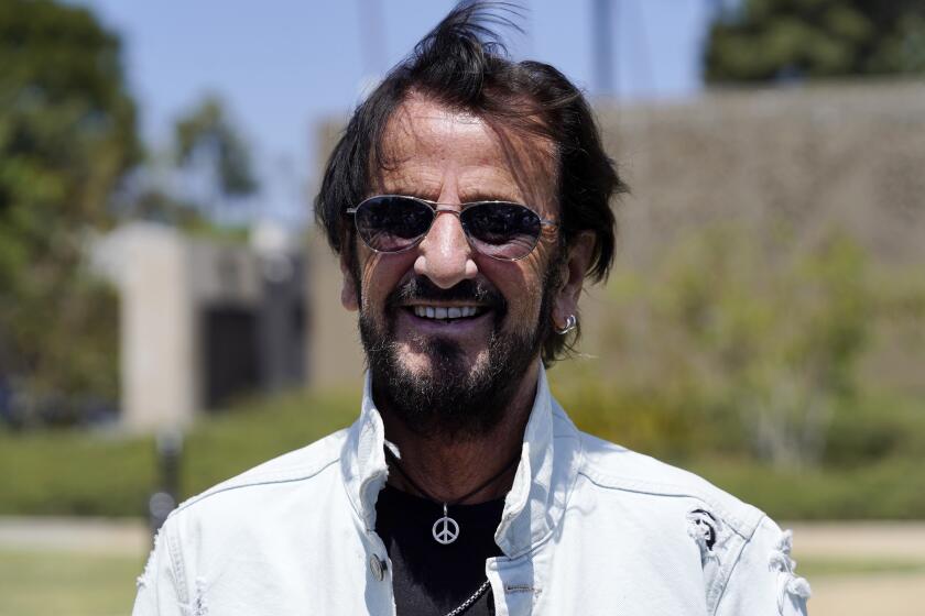A man wearing sunglasses and a white jacket smiles outdoors